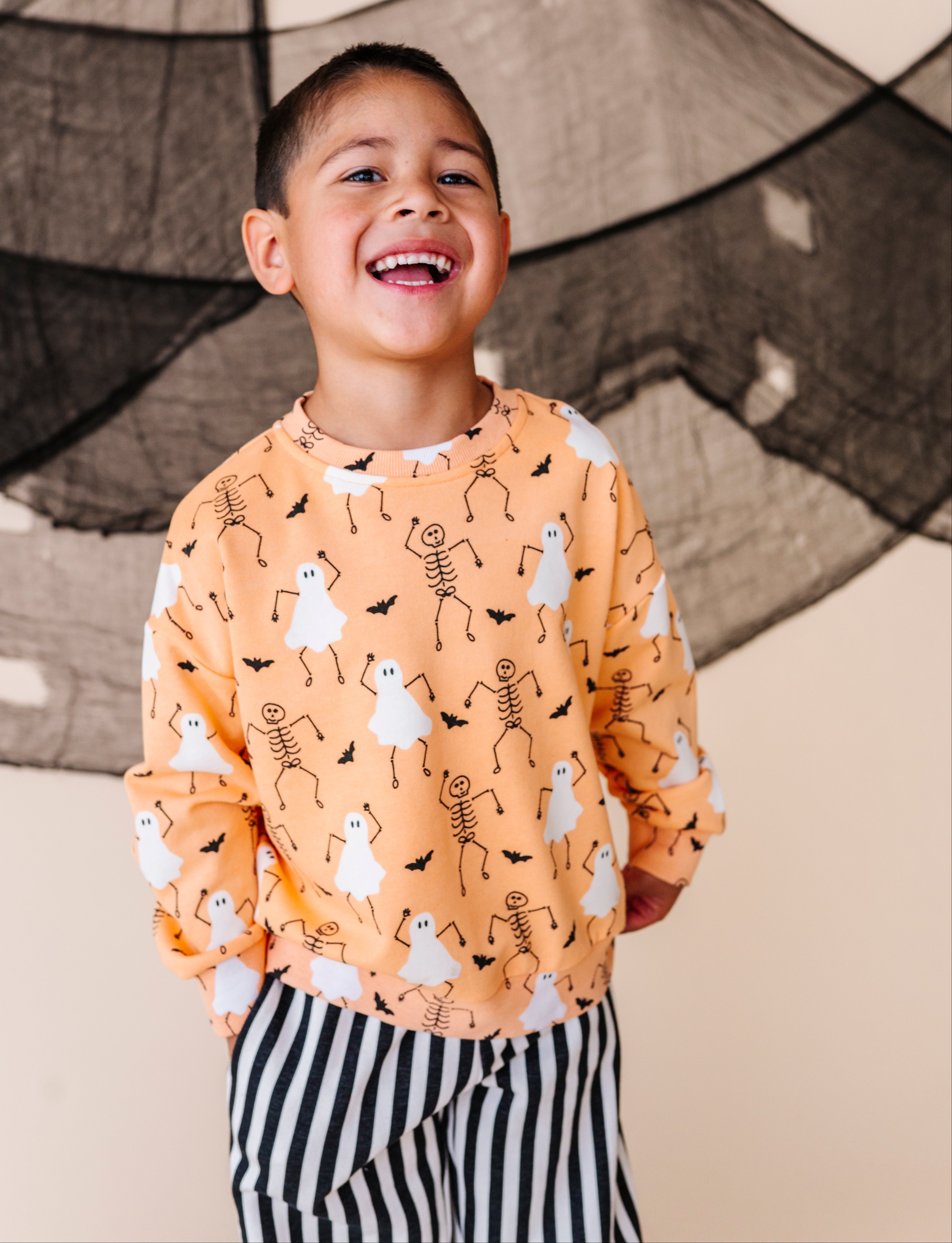 Child wearing a Halloween-themed sweatshirt with ghosts, skeletons, and bats against a decorative backdrop.